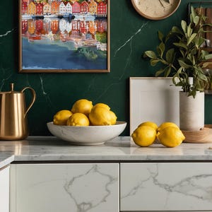May include: A kitchen scene featuring a watercolor painting of a colorful waterfront town, a gold pitcher, a marble bowl filled with lemons, and a wooden clock. The countertop and cabinets have a marble design, and the wall is a dark green color.