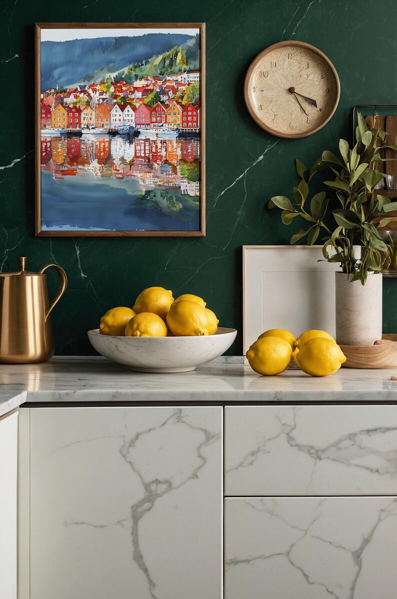 May include: A kitchen scene featuring a watercolor painting of a colorful waterfront town, a gold pitcher, a marble bowl filled with lemons, and a wooden clock. The countertop and cabinets have a marble design, and the wall is a dark green color.