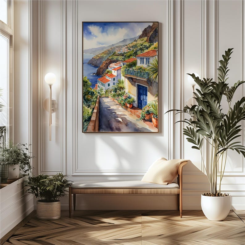 May include: Watercolor painting of a white building with a blue door on a hillside overlooking the ocean. The building is surrounded by lush greenery and flowers. The painting is framed and hanging on a white wall.