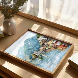 Peut inclure: Une impression encadr&eacute;e &agrave; l'aquarelle repr&eacute;sentant un village au bord d'un lac avec une montagne en arri&egrave;re-plan. L'&oelig;uvre pr&eacute;sente des b&acirc;timents aux toits rouges et oranges, un clocher d'&eacute;glise et un reflet dans l'eau. Le cadre est en bois clair.