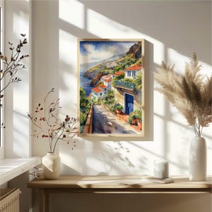 May include: Watercolor painting of a white building with a blue door and red tile roof on a hillside overlooking the ocean. The building is surrounded by lush greenery and flowers. The painting is framed in a light wood frame.