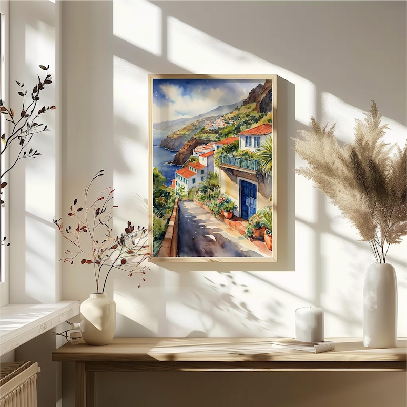 May include: Watercolor painting of a white building with a blue door and red tile roof on a hillside overlooking the ocean. The building is surrounded by lush greenery and flowers. The painting is framed in a light wood frame.