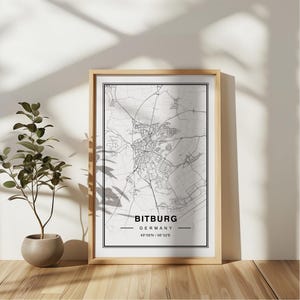 May include: A black and white map print of Bitburg, Germany. The map is framed in a light wood frame and is hanging on a white wall. The text "Bitburg, Germany 49°58'N / 6°32'E" is printed below the map.