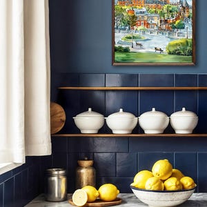 May include: A framed watercolor painting of a European cityscape hangs on a navy blue wall. Below, white ceramic bowls with lids sit on a wooden shelf. A bowl of lemons and a wooden cutting board are on the countertop.