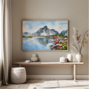 May include: Watercolor painting of a coastal village with red buildings, nestled at the base of a mountain range. The artwork is framed and hangs above a wooden bench. The water reflects the sky and mountains.