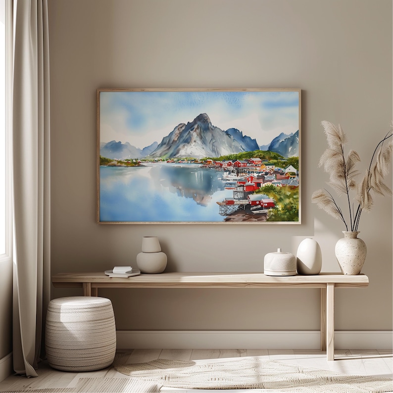 May include: Watercolor painting of a coastal village with red buildings, nestled at the base of a mountain range. The artwork is framed and hangs above a wooden bench. The water reflects the sky and mountains.