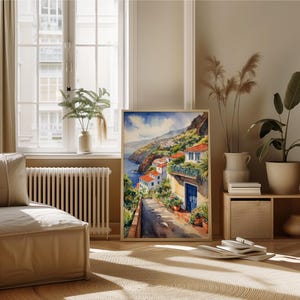 May include: Watercolor painting of a coastal town with white buildings and red tile roofs. The town is nestled on a hillside overlooking a blue ocean. The painting is framed in a light wood frame.