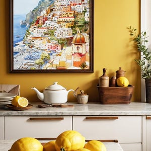May include: A watercolor painting of a coastal Italian town hangs above a kitchen counter. A white teapot, lemons, and a wooden bowl filled with lemons are on the counter. The painting depicts colorful buildings on a hillside overlooking the sea.
