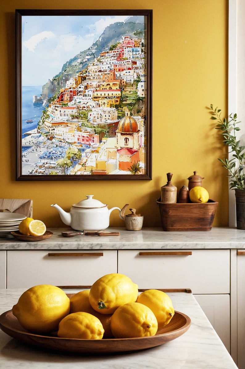 May include: A watercolor painting of a coastal Italian town hangs above a kitchen counter. A white teapot, lemons, and a wooden bowl filled with lemons are on the counter. The painting depicts colorful buildings on a hillside overlooking the sea.