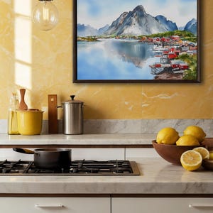 May include: A framed watercolor painting depicts a coastal village with red buildings nestled at the base of a mountain range. The artwork is displayed above a kitchen countertop with lemons in a wooden bowl.