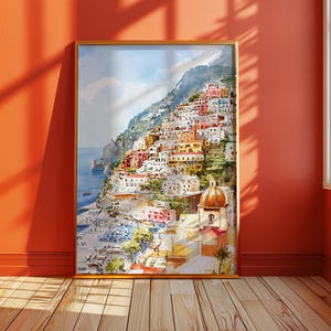 May include: A framed watercolor painting of a coastal Italian town. The artwork depicts colorful buildings cascading down a hillside towards the sea. The painting features a warm color palette with shades of orange, red, yellow, and blue.