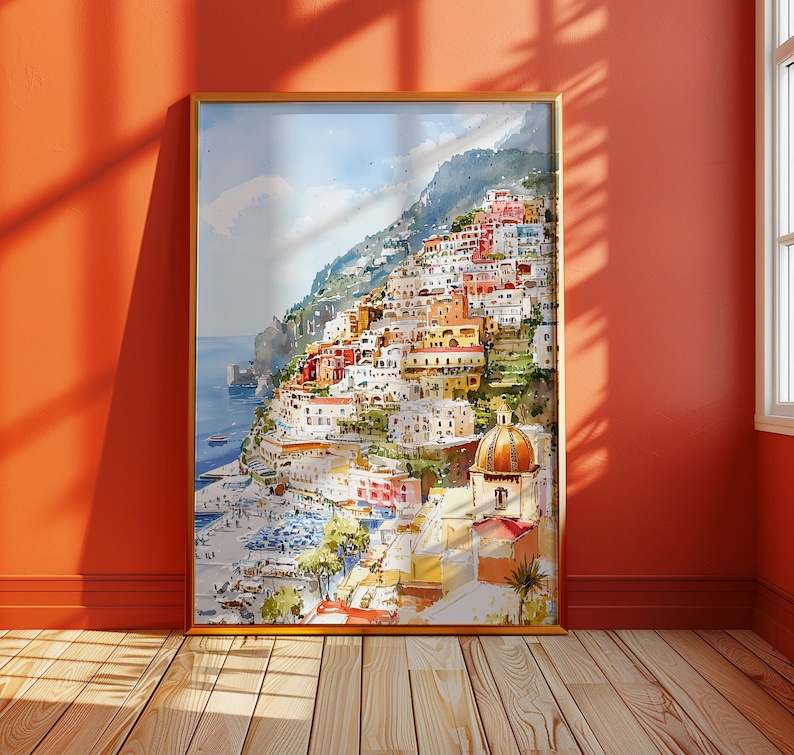 May include: A framed watercolor painting of a coastal Italian town. The artwork depicts colorful buildings cascading down a hillside towards the sea. The painting features a warm color palette with shades of orange, red, yellow, and blue.