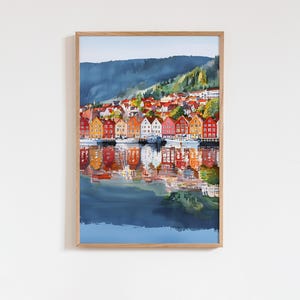 May include: Watercolor print of a coastal town with colorful buildings reflected in the water. The artwork features a mountain in the background and a wooden frame.