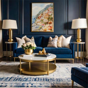 May include: A luxurious living room featuring a blue velvet sofa, gold accent tables with lamps, and a round coffee table. A framed watercolor painting of a coastal town hangs above the sofa. Decorative pillows and a floral arrangement add to the room's elegance.