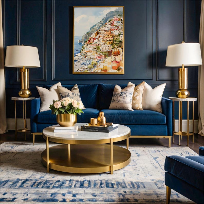 May include: A luxurious living room featuring a blue velvet sofa, gold accent tables with lamps, and a round coffee table. A framed watercolor painting of a coastal town hangs above the sofa. Decorative pillows and a floral arrangement add to the room's elegance.