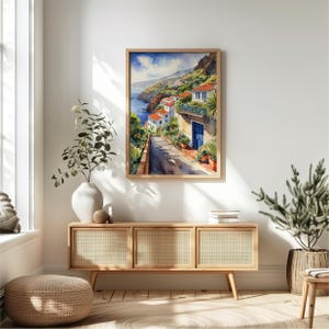 May include: A watercolor painting of a coastal town with white buildings and a blue door. The painting is framed in a light wood frame and is hanging on a white wall.