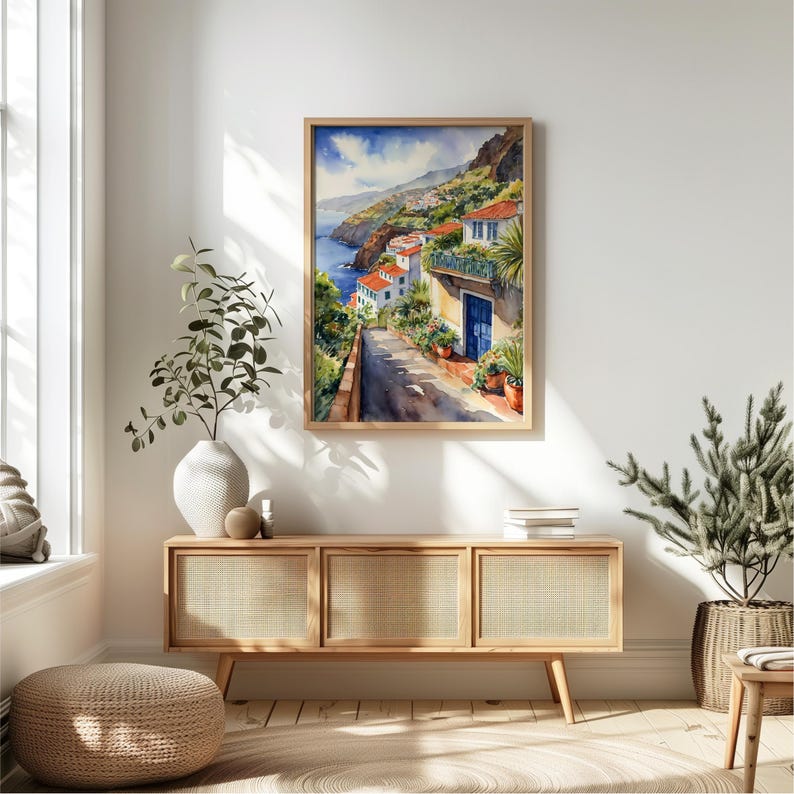 May include: A watercolor painting of a coastal town with white buildings and a blue door. The painting is framed in a light wood frame and is hanging on a white wall.