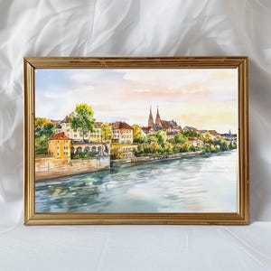 May include: Watercolor painting of a European cityscape in a gold frame. The artwork depicts buildings along a river, with a church in the background. The sky is a mix of blue, pink, and yellow.