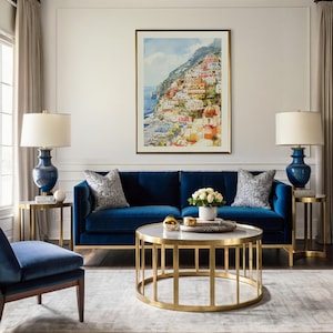 May include: A living room scene featuring a navy blue velvet sofa, a round gold coffee table with a marble top, and two matching side tables. A framed watercolor painting of a coastal town hangs above the sofa. Two navy blue lamps with white shades are on the side tables.