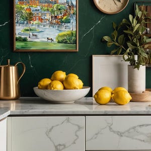 May include: A kitchen scene featuring a watercolor painting of a cityscape in a wooden frame, a gold pitcher, a white marble bowl filled with lemons, and a wooden clock. The background is a dark green marble wall.