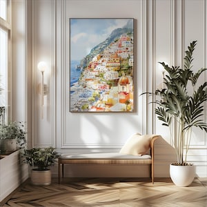 May include: A framed watercolor painting depicts a colorful coastal town nestled on a hillside. The artwork features buildings in shades of red, orange, and white, with a blue sea and sky. The painting hangs above a wooden bench with a cream-colored cushion.