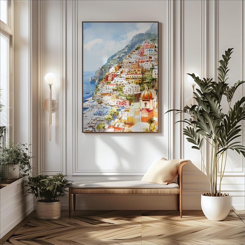 May include: A framed watercolor painting depicts a colorful coastal town nestled on a hillside. The artwork features buildings in shades of red, orange, and white, with a blue sea and sky. The painting hangs above a wooden bench with a cream-colored cushion.