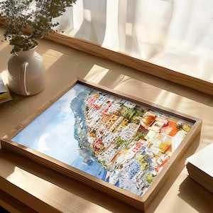 May include: A framed watercolor print of a coastal Italian town. The artwork depicts colorful buildings cascading down a hillside towards the sea. The frame is made of light-colored wood, and the print is displayed on a wooden table.