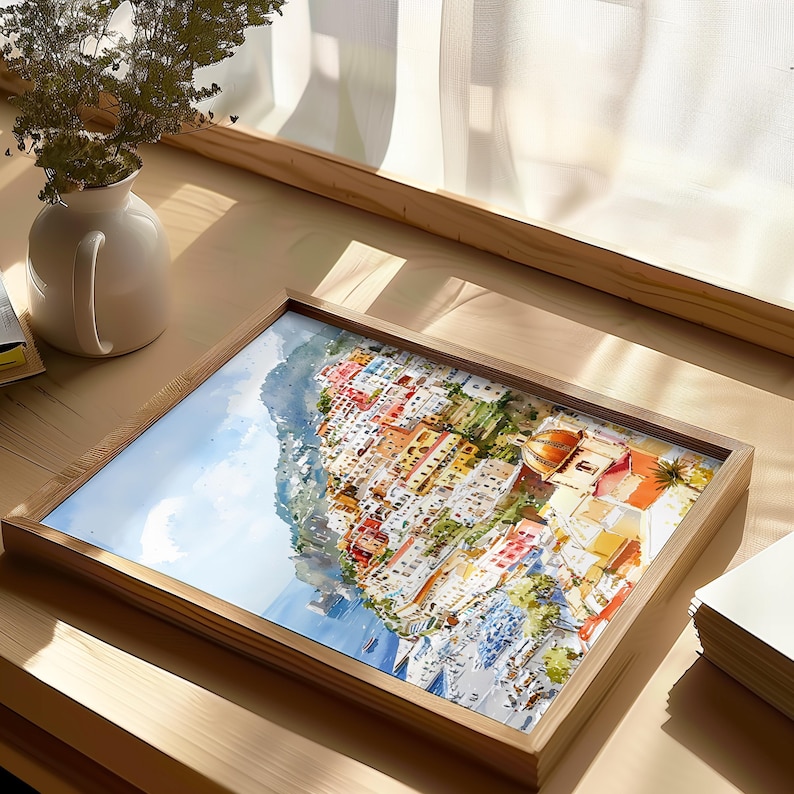 May include: A framed watercolor print of a coastal Italian town. The artwork depicts colorful buildings cascading down a hillside towards the sea. The frame is made of light-colored wood, and the print is displayed on a wooden table.