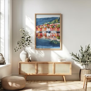 May include: A framed watercolor print depicts a colorful waterfront scene with buildings reflected in the water. The artwork features a variety of warm colors, including red, orange, and yellow, set against a backdrop of a mountain and blue sky.