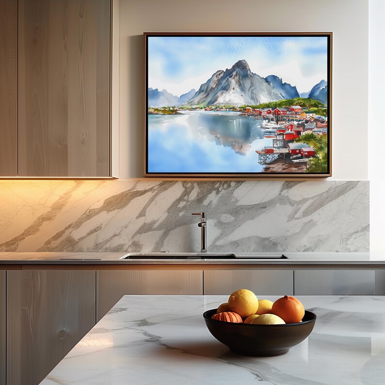 May include: A framed watercolor painting depicts a mountain landscape with a lake and village. The artwork features a mountain range, a body of water, and red-roofed buildings. The painting is displayed above a kitchen countertop with a bowl of fruit.