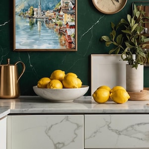 Peut inclure: Int&eacute;rieur de cuisine avec un tableau encadr&eacute; repr&eacute;sentant un paysage &agrave; l'aquarelle, une carafe dor&eacute;e et un bol de citrons. Une horloge ronde est accroch&eacute;e au mur en marbre vert fonc&eacute;. Le plan de travail est en marbre blanc.