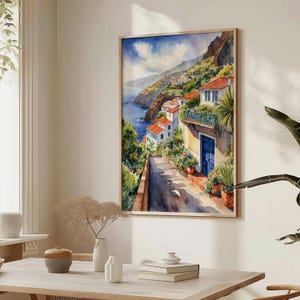 May include: Watercolor painting of a colorful hillside village with white houses and a blue door. The village is situated on a cliff overlooking a blue ocean. The painting is framed in a light wood frame.