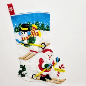 May include: A white felt Christmas stocking featuring a blue sky background with green pine trees. A snowman in a black top hat and a Santa Claus are skiing on the stocking. The stocking includes a red hanging loop and a yellow sign reading NORTH POLE.
