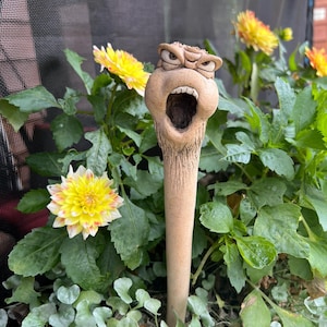 May include: A tan, cylindrical garden stake featuring a sculpted, expressive face at the top. The figure has a wide, open mouth, furrowed brows, and visible teeth, with a textured, earthy finish that tapers into a long, smooth, narrow stem.