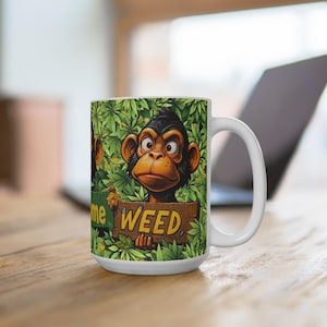 May include: White ceramic mug featuring a cartoon monkey holding a wooden sign that reads "WEED". The mug has a green background of cannabis leaves. The monkey is brown and black with a surprised expression.