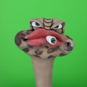 May include: A sculpted clay figure of a snake head with a tan and brown spotted pattern. The figure features prominent red lips, narrow grey eyes, and a small white cigarette-like object held between the lips. The sculpture has a long neck.