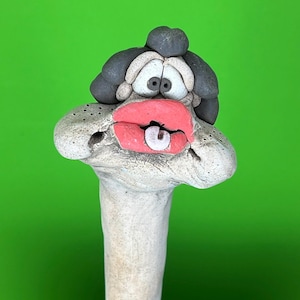 May include: A hand-sculpted clay figure featuring a long, pale, textured neck and a head with dark gray hair. The face has wide, round eyes, prominent bright red lips, and a small white tongue sticking out, creating a whimsical, expressive character.