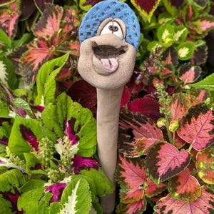 May include: A clay garden stake featuring a sculpted, light brown creature with a wide, open mouth, a protruding pink tongue, and large eyes. The creature wears a textured, bright blue cap on its head, and the entire figure is mounted on a long, slender, cylindrical clay stem.