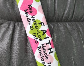 May include: A fabric seatbelt cover with a pattern of pink hearts and green circles on a white background. The cover has the words "FAITH" and "Non Verbal-Low Medical Info Muscle Tone Enclosed" printed on it in black.
