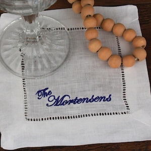 May include: A white linen cocktail napkin with a decorative border and the embroidered name "The Mortensens" in blue script. A clear glass sits on the napkin, and a strand of wooden beads rests nearby.