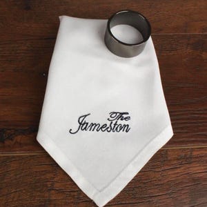 May include: A white linen napkin with the embroidered text "The Jameston" in black script. A dark gray metal napkin ring sits at the top. The napkin is folded and placed on a wooden surface.