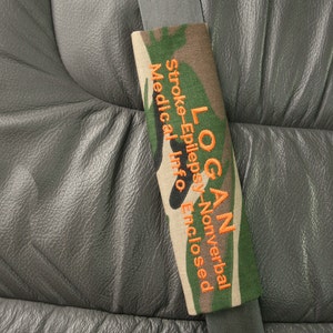 May include: Camouflage seatbelt cover with the text "LOGAN Stroke-Epilepsy-Nonverbal Medical Info Enclosed" embroidered in orange.