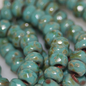 May include: Strands of turquoise ceramic beads with a speckled, aged appearance. The beads are round with a slightly flattened shape and a glossy finish. The beads are strung together, creating a necklace or bracelet.