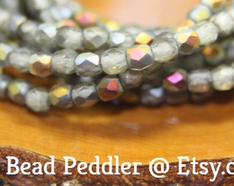 Op de afbeelding: Een close-up van een streng iriserende grijze glazen kralen. De kralen zijn gefacetteerd en hebben een subtiele glans. De kralen zijn in een cirkelvormige patroon gerangschikt, wat een prachtig en elegant ontwerp creëert.