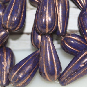 Puede incluir: Una hebra de cuentas en forma de lágrima con un esmalte morado oscuro y detalles dorados. Las cuentas tienen una superficie texturizada y están enhebradas en un hilo transparente.
