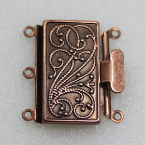 May include: A copper-toned metal clasp with a decorative filigree design. The clasp has three loops on each side for attaching jewellery.