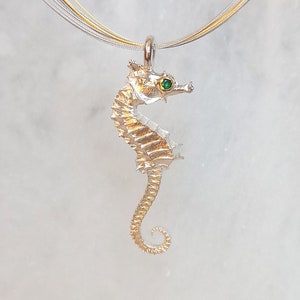 Op de afbeelding: Een gouden zeepaardje hanger met een groene edelsteen oog, hangend aan een zilveren ketting.