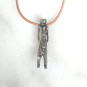 May include: A silver pendant necklace featuring a detailed, abstract human figure. The pendant is attached to a brown leather cord.