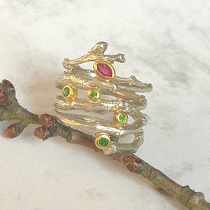 Peut inclure: Bague argentée au design inspiré de branches, ornée d'un rubis en forme de marquise et de plusieurs pierres vertes rondes serties d'or. La bague est présentée sur une petite branche, mettant en valeur ses détails.