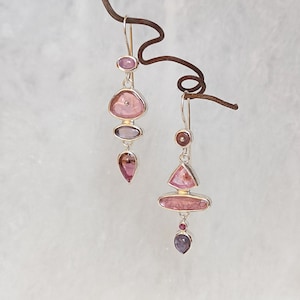 Könnte beinhalten: Zwei silberne Ohrringe mit rosa und lila Edelsteinen. Jeder Ohrring hat eine einzigartige Anordnung von Formen, darunter Ovale, Dreiecke und Tropfen. Die Edelsteine sind in Silber gefasst, was ein zartes und elegantes Design ergibt.